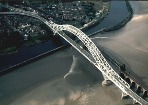 The Mersey Bore passing the Runcorn road and railway bridges, following the main channel (right to left) just before reaching the wall of the Manchester Ship Canal where it gets reflected.